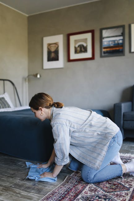 A woman with brown hair tied back, dressed in a light blue striped shirt and blue jeans, is shown kneeling on a traditional patterned rug in a bedroom. She is demonstrating surface cleaning by wiping a wooden floor with a blue cloth. The room features a bed with grey bedding and a metal frame, positioned against a light-colored wall decorated with three framed pictures. A blue armchair is visible in the corner, and natural light illuminates the space. The scene reflects domestic cleaning and maintenance activities, highlighting cleanliness and attention to detail. Streatham Cleaners offers professional deep cleaning services for homes, ensuring hygiene and surface sanitation.