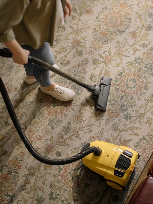 A person operating a yellow portable fan or air scrubber on a patterned carpet in a room with wooden flooring. The individual is holding the device with both hands, focusing on the surface cleaning process for deep cleaning or sanitisation as part of a domestic cleaning service. The carpet features a traditional floral or oriental design in muted tones, and the room is softly lit, emphasizing cleanliness and hygiene. The scene highlights professional cleaning routines offered by Streatham Cleaners in their residential carpet cleaning service, maintaining high standards of surface cleanliness and hygiene.
