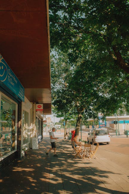 The image depicts a sidewalk scene outside a shop on Streatham High Road, with the store's blue sign partially visible. The sidewalk is shaded by a large leafy tree, casting dappled sunlight and shadows on the pavement. The shop exterior features a wooden overhang and a glass storefront, with a concrete or stone surface outside. A woman in casual clothing is walking along the sidewalk, while another person is seated at an outdoor seating area consisting of wooden tables and chairs. Several parked cars are visible further down the street. The scene suggests a clean, well-maintained environment suitable for pedestrian and customer activity, aligning with the kind of surface and area that Streatham Cleaners might service for local businesses, ensuring hygienic, well-maintained premises.