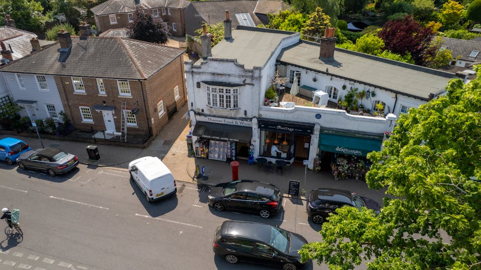 An aerial view of a commercial street with small shops on Streatham High Road, showing clean and well-maintained building facades. One shop has a white exterior with large display windows and a black awning, while another has a green awning with the sign 'SRETT GARDEN'. The pavement is clear and tidy, with parked cars along the curb and a few traffic cones. Trees with lush green leaves line the street, providing shade. The scene is bright, with natural sunlight highlighting the surfaces of the buildings and street, reflecting a neat and hygienic urban environment. Streatham Cleaners offers surface cleaning and deep sanitisation services, ensuring commercial premises like these maintain high standards of hygiene and cleanliness.