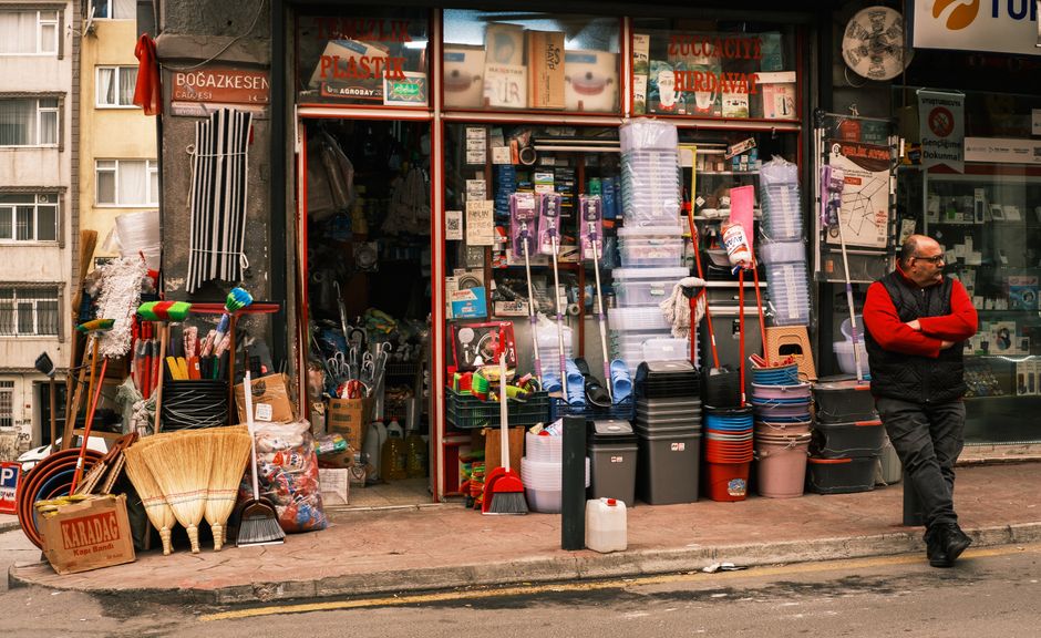 The image depicts a sidewalk scene outside a shop on Streatham High Road, with the store's blue sign partially visible. The sidewalk is shaded by a large leafy tree, casting dappled sunlight and shadows on the pavement. The shop exterior features a wooden overhang and a glass storefront, with a concrete or stone surface outside. A woman in casual clothing is walking along the sidewalk, while another person is seated at an outdoor seating area consisting of wooden tables and chairs. Several parked cars are visible further down the street. The scene suggests a clean, well-maintained environment suitable for pedestrian and customer activity, aligning with the kind of surface and area that Streatham Cleaners might service for local businesses, ensuring hygienic, well-maintained premises.