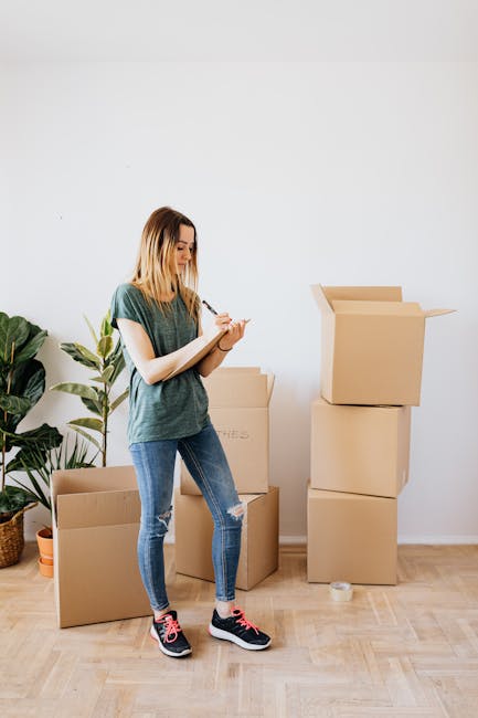 A young woman with blonde hair, dressed in a casual green T-shirt, ripped blue jeans, and black sneakers with pink accents, stands on a light wooden floor in a bright room with white walls. She is taking notes with a pen and notepad. Behind her, there are several cardboard moving boxes, some stacked and one partially open, indicating an end of tenancy or moving process. To her left, a large green potted plant is placed on the floor, adding a touch of natural decor to the space. The scene is well-lit with natural light, emphasizing the cleanliness and neatness of the room, which suggests it is prepared for professional surface cleaning or deep cleaning services offered by Streatham Cleaners, as referenced on the Streatham Hill end of tenancy cleaning checklist webpage.