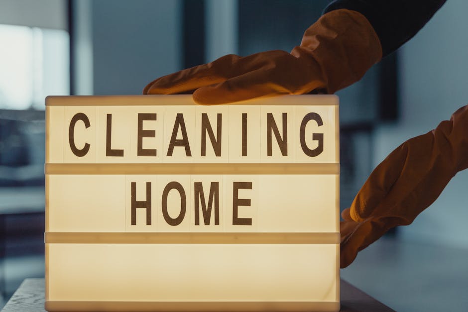 A person wearing brown cleaning gloves is placing a lightbox sign that reads 'CLEANING HOME' indoors. The sign is illuminated from within, with black capital letters on a white background. The setting