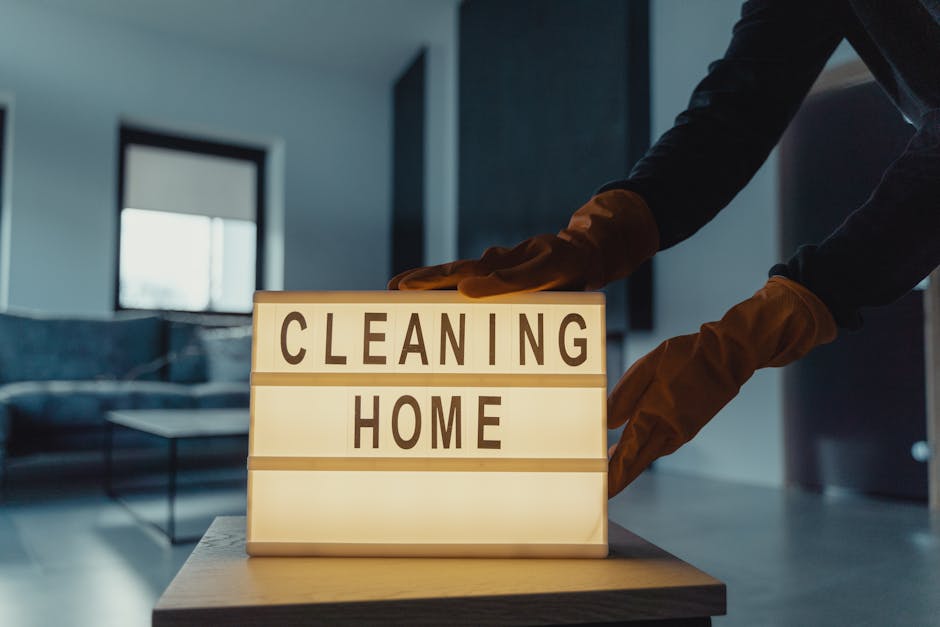 A person wearing brown cleaning gloves is placing a lightbox sign that reads 'CLEANING HOME' indoors. The sign is illuminated from within, with black capital letters on a white background. The setting